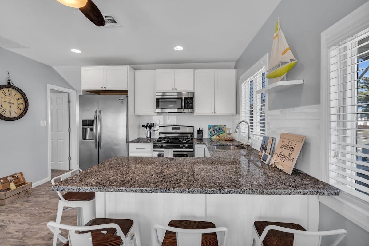 A modern kitchen area is showcased, featuring sleek white cabinetry and granite countertops. Stainless steel appliances, including a refrigerator and stove, are visible. A central island with seating is flanked by several white chairs, and natural light filters through the windowed walls.