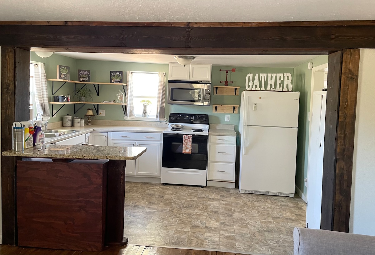 The kitchen area is designed with white cabinetry and a light granite countertop. Shelves display decorative items. A black microwave and stove are positioned against the wall, accompanied by a white refrigerator. Natural light enters through the window above the sink, creating a bright atmosphere.