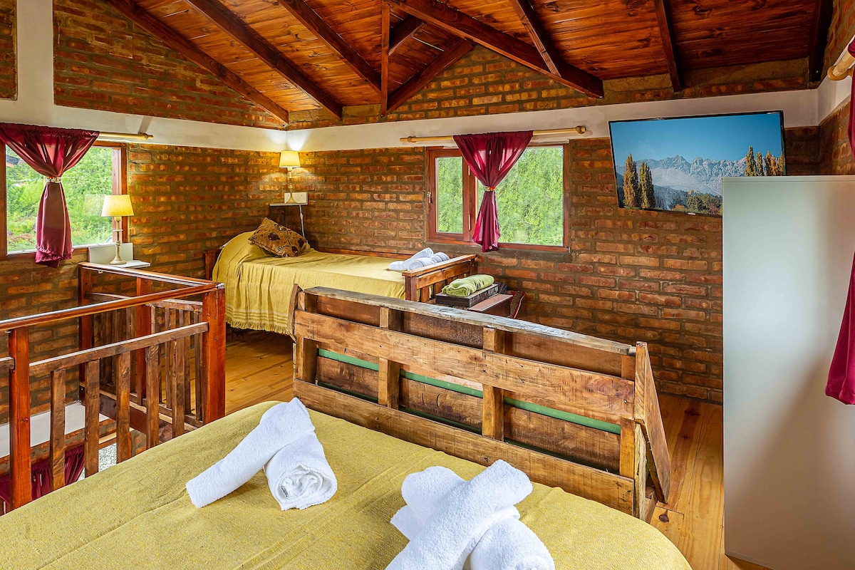An inviting sleeping area features two beds, each adorned with fresh towels. Exposed wooden beams complement the brick walls, while large windows allow natural light to fill the room. A television is mounted on one wall, enhancing the cozy atmosphere.
