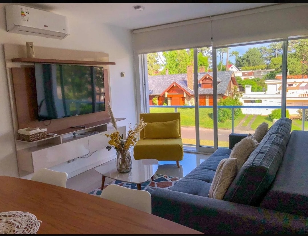 A modern living area features a comfortable gray sofa and a vibrant yellow chair, arranged around a glass coffee table. A large television is mounted on the wall opposite the seating area. Floor-to-ceiling windows allow natural light to fill the space, with serene outdoor views visible.