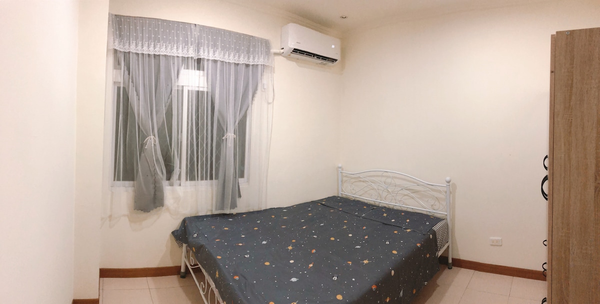 A simple bedroom features a spacious bed with a star-patterned blanket. Natural light enters through neatly draped curtains over a window, enhancing the room's bright and airy feel. An air conditioning unit is mounted on the wall to provide comfort.