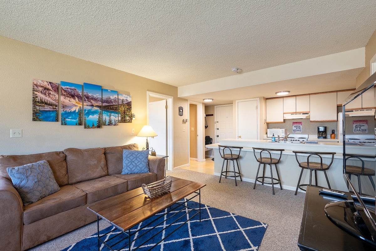 A welcoming living area showcases a large brown sofa adorned with decorative pillows. A coffee table sits in front, complemented by a blue area rug. The space is decorated with landscape artwork, and a kitchen bar with stools is visible in the background.