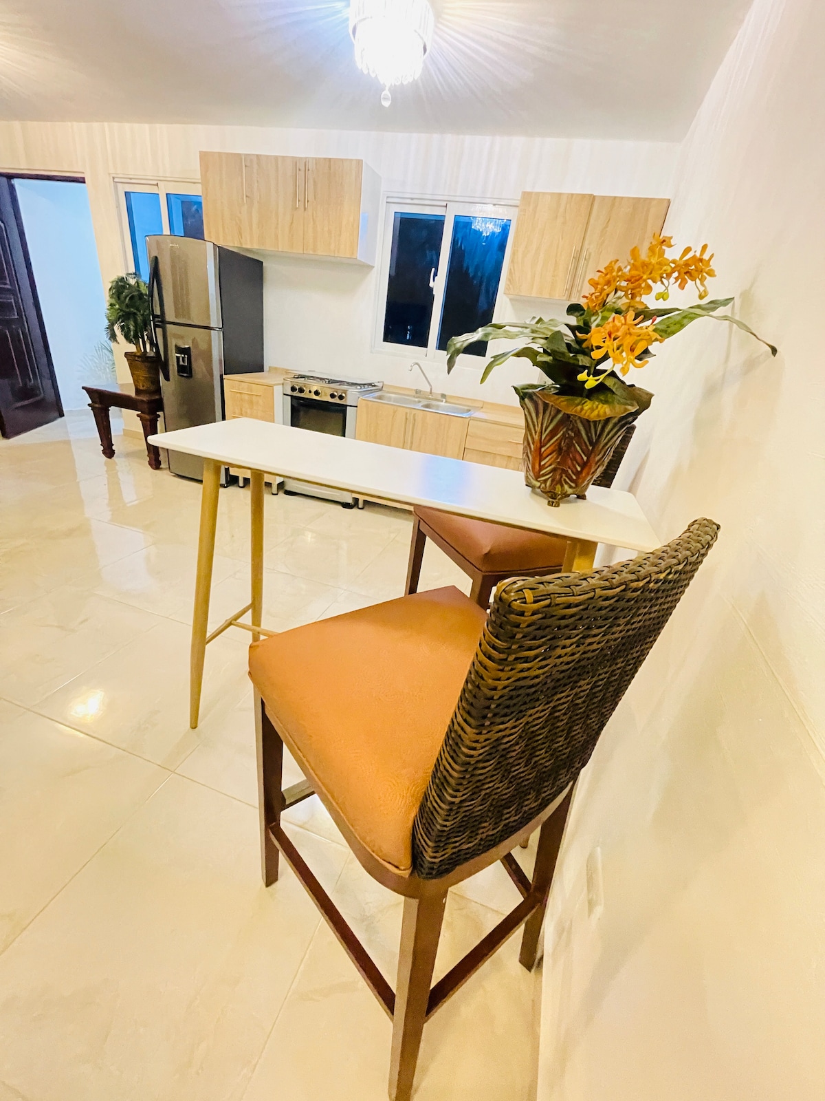 A modern kitchen area features a sleek bar table paired with two tall chairs. One chair has a woven design, while the other is upholstered in a warm orange hue. A decorative vase filled with yellow flowers adds a touch of color next to the seating.