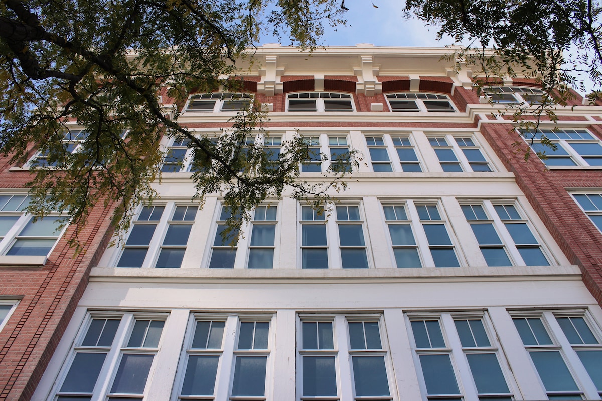 The historic building is showcased from a low angle, highlighting its striking red brick façade and large, evenly spaced windows. Trees in the foreground provide a natural frame, while the blue sky is visible above, contributing to the urban character of the location.