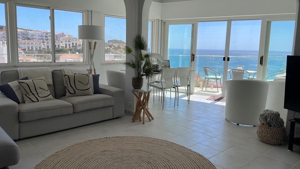 A light-filled living area features large glass windows offering ocean views. A comfortable sofa with decorative pillows is positioned beside a round area rug. A dining set for six is visible in the background, complemented by a white armchair and potted plant.