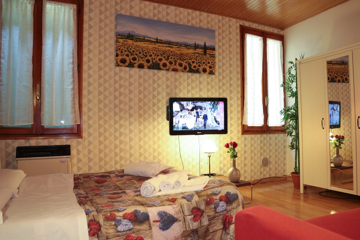 A cozy sleeping area features a patterned bedspread with rolled towels arranged neatly on the bed. A television is mounted on the wall, complemented by a floor lamp and a small plant nearby. Large windows with sheer curtains allow natural light to fill the space.