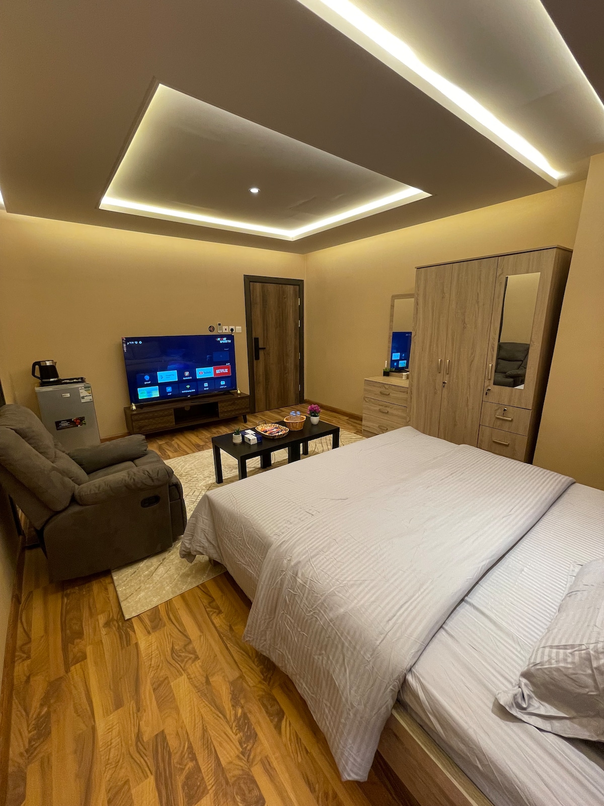 A modern room is displayed with warm lighting and wooden flooring. The spacious bed is neatly made with light-colored linens. A comfortable armchair and a television are positioned near a coffee table featuring snacks. A wardrobe with mirrored doors adds functionality to the space.