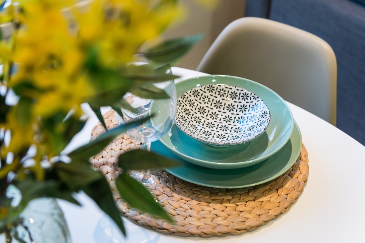 A dining setting is presented, featuring a round white table adorned with a woven placemat. Two stacked plates, one in a soft green shade and the other decorated with a subtle floral pattern, are complemented by a glass and a vase with yellow flowers in the background.