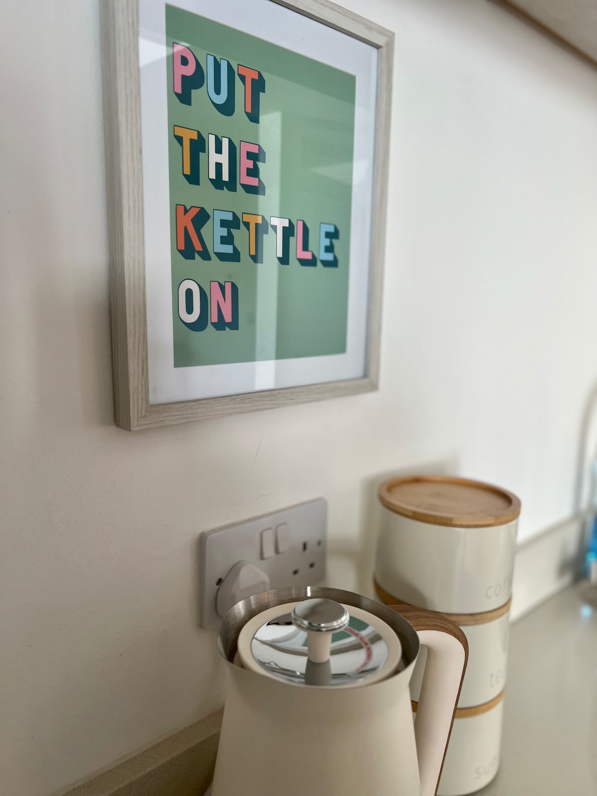 A modern kitchen corner displays a sleek electric kettle beside decorative storage containers. A framed artwork with colorful letters reads 'PUT THE KETTLE ON'. Light reflects softly off the surfaces, providing a fresh and inviting atmosphere.