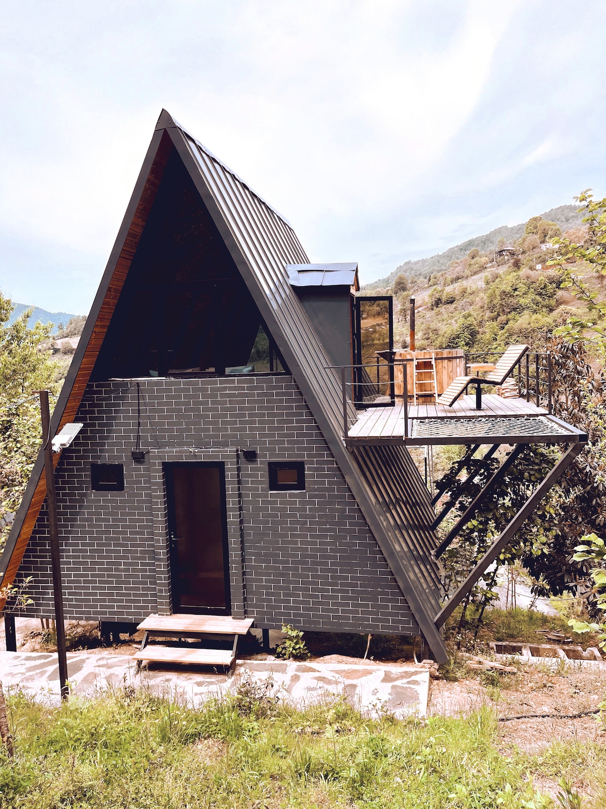 A modern triangular structure is framed by lush greenery, showcasing a combination of dark brick and sleek metal exterior. A wooden deck with seating is present on the upper level, enhancing the outdoor living space while large windows allow ample natural light.