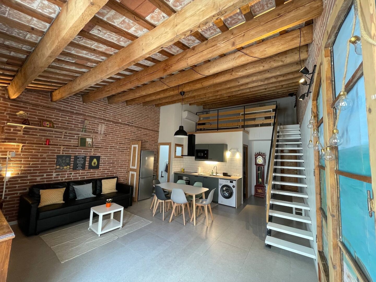 The spacious living area features exposed wooden beams and a blend of brick walls. A black sofa is paired with a modern dining table surrounded by chairs. An open kitchen area includes sleek appliances. A wooden staircase leads to a loft area above.
