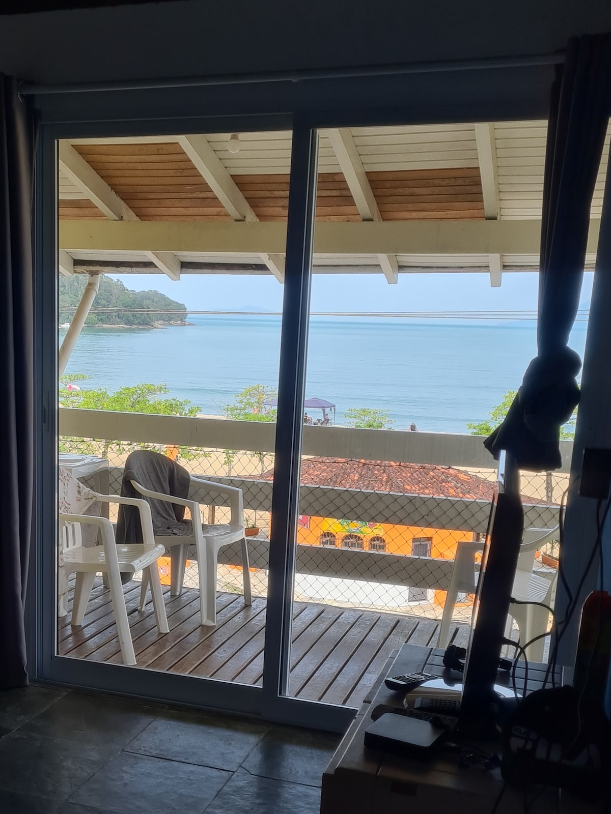 A spacious balcony is seen through a glass sliding door, offering a view of the calm sea. Two white chairs are positioned on the wooden deck, and colorful buildings are visible in the distance, framed by lush greenery and blue skies.