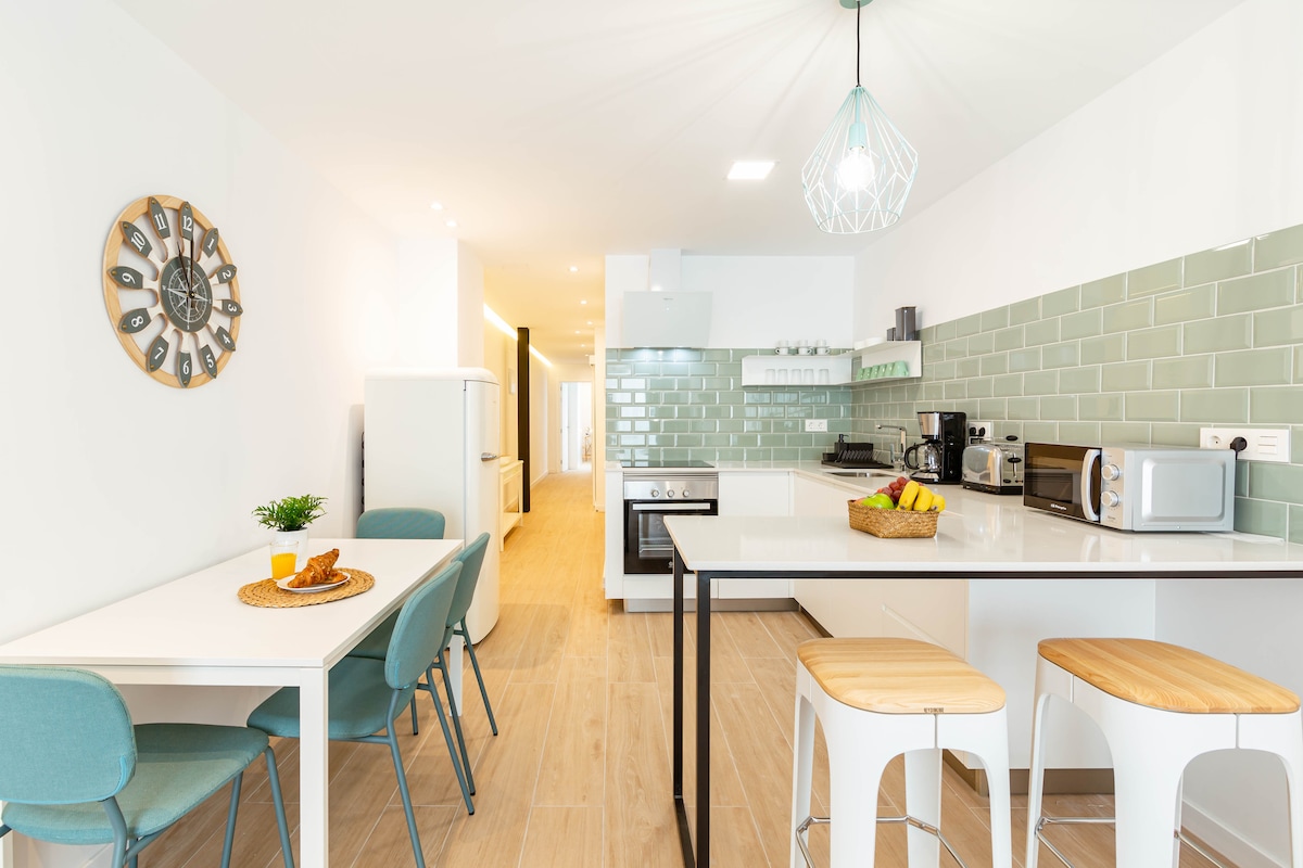 A clean and modern kitchen area is presented with light wood flooring. A table with turquoise chairs invites dining, while a fruit bowl adds a touch of color. Equipped appliances, including a refrigerator and microwave, are visible against a backdrop of green tile backsplash.