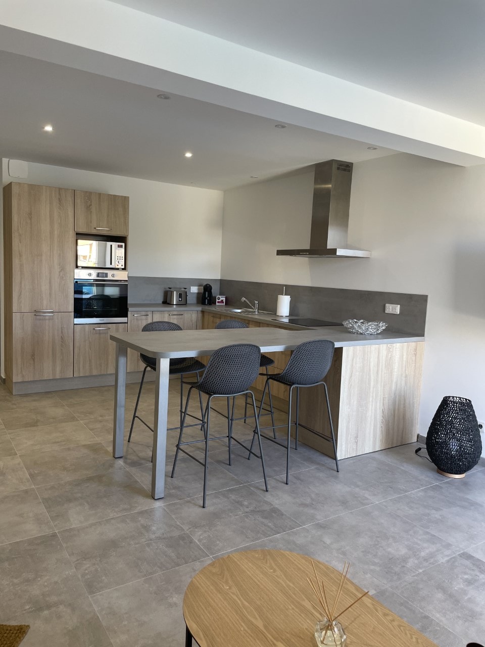 A modern kitchen is presented, featuring light wood cabinetry and a spacious countertop. Four bar stools are arranged at the countertop, complemented by stainless steel appliances. The floor is tiled in a neutral grey, contributing to a clean and functional cooking space.