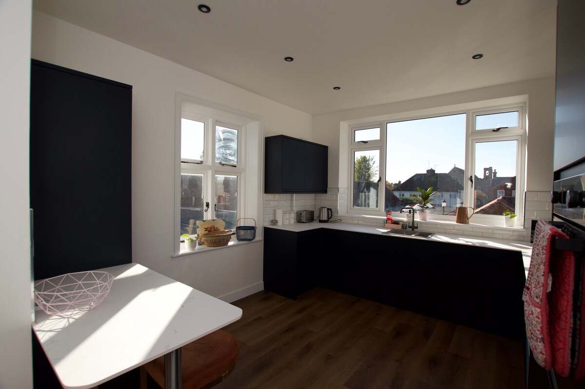 A modern kitchen features blue cabinetry and a white countertop. Natural light fills the space through multiple windows, highlighting the minimalist design. A small table with a round chair is positioned near the window, complemented by appliances neatly arranged along the counter.