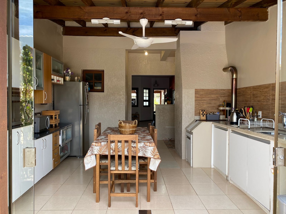 An open kitchen and dining area features wood-beamed ceilings and tile flooring. A wooden table is set with a basket, surrounded by matching chairs. Stainless steel appliances and well-organized countertops offer ample space for cooking and dining. Natural light pours in through large glass doors.