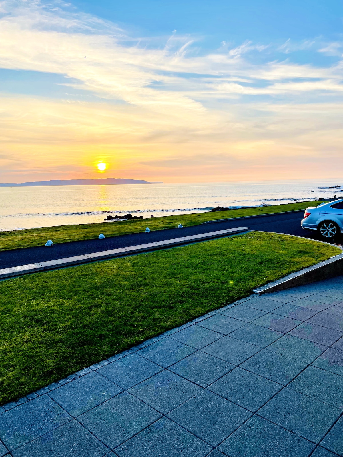 A serene coastal view at sunset reveals the shimmer of the ocean under a soft orange sky. A lawn with fresh green grass borders the scene, while a paved pathway leads towards the water's edge. A parked car is positioned nearby, offering a perspective of the tranquil landscape.