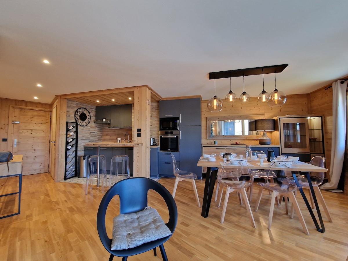 An open-plan living area is presented with warm wooden accents. A modern kitchen space features high-end appliances and a clock mounted on the wall. A dining table with clear chairs is positioned nearby, creating a comfortable space for meals and gatherings.