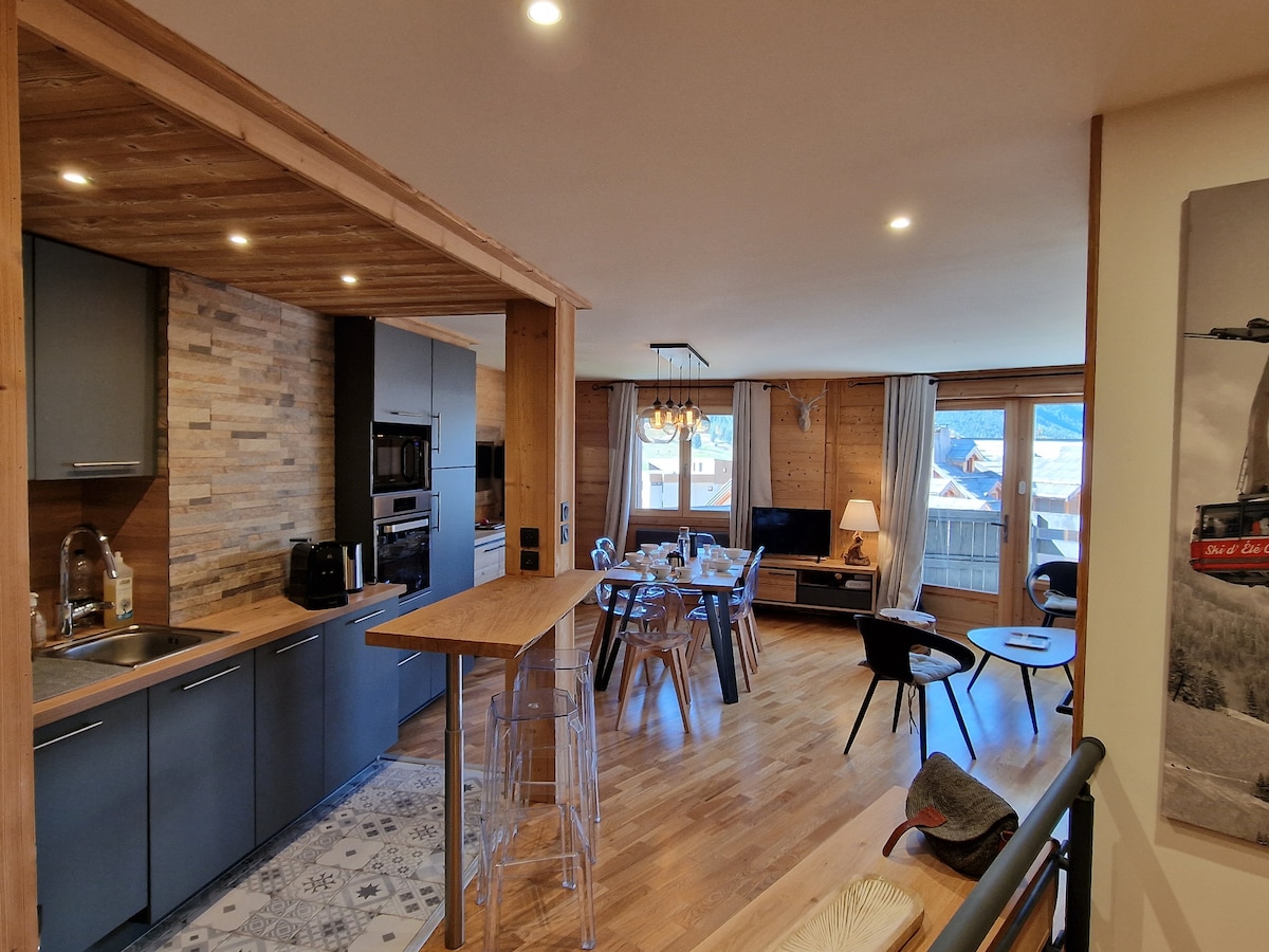 The open-concept living area features a modern kitchen with sleek cabinets and a countertop, complemented by a dining table surrounded by clear chairs. Natural light fills the space through large windows, showcasing mountain views. A cozy seating area includes a TV and decorative elements.
