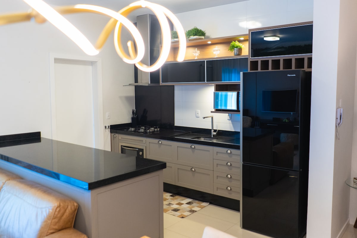A modern kitchen is equipped with sleek black appliances and a spacious granite countertop. Stylish cabinetry features a mix of dark and light tones, while decorative plants add subtle accents. The layout includes essential cooking amenities, ensuring a functional space for meal preparation.