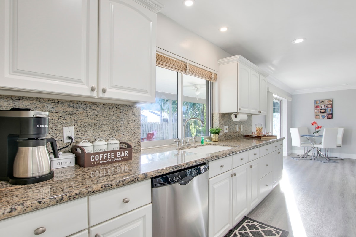 A modern kitchen features white cabinetry and a spacious countertop made of light granite. A coffee maker and a coffee bar set are positioned on the counter. Sunlight pours in through a large window, illuminating the room, which connects to a dining area with a small table and chairs.