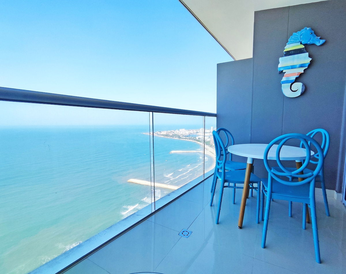 A balcony area features a round table surrounded by four blue chairs, overlooking a panoramic view of the ocean. The clear blue sky complements the serene coastal landscape, while a colorful seahorse wall decoration adds a touch of charm to the space.