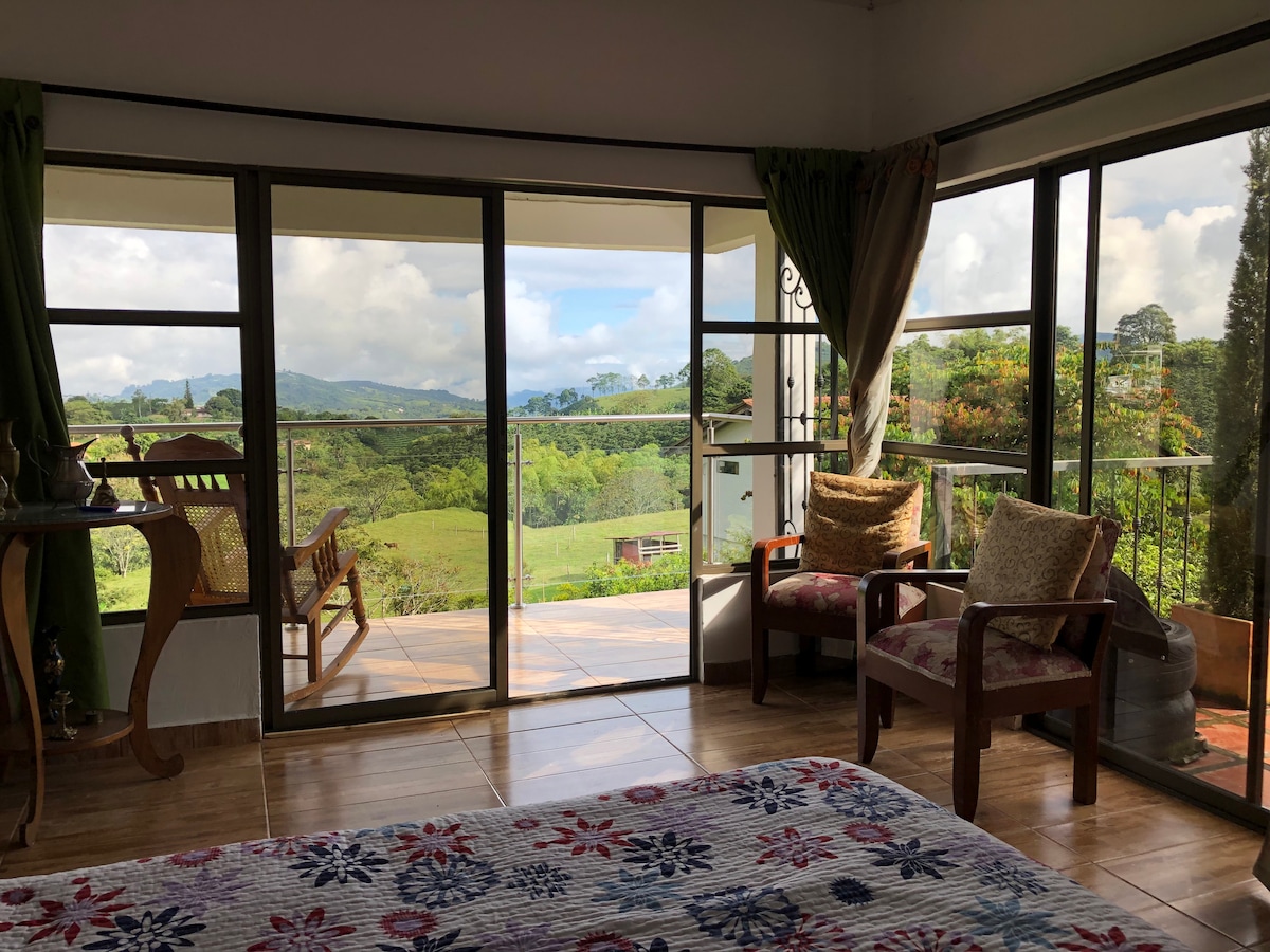 Large glass doors provide a view of the lush green landscape beyond. A comfortable seating area with two armchairs sits next to the doors, while a spacious bed with a colorful patterned blanket occupies the foreground. Natural light floods the room.