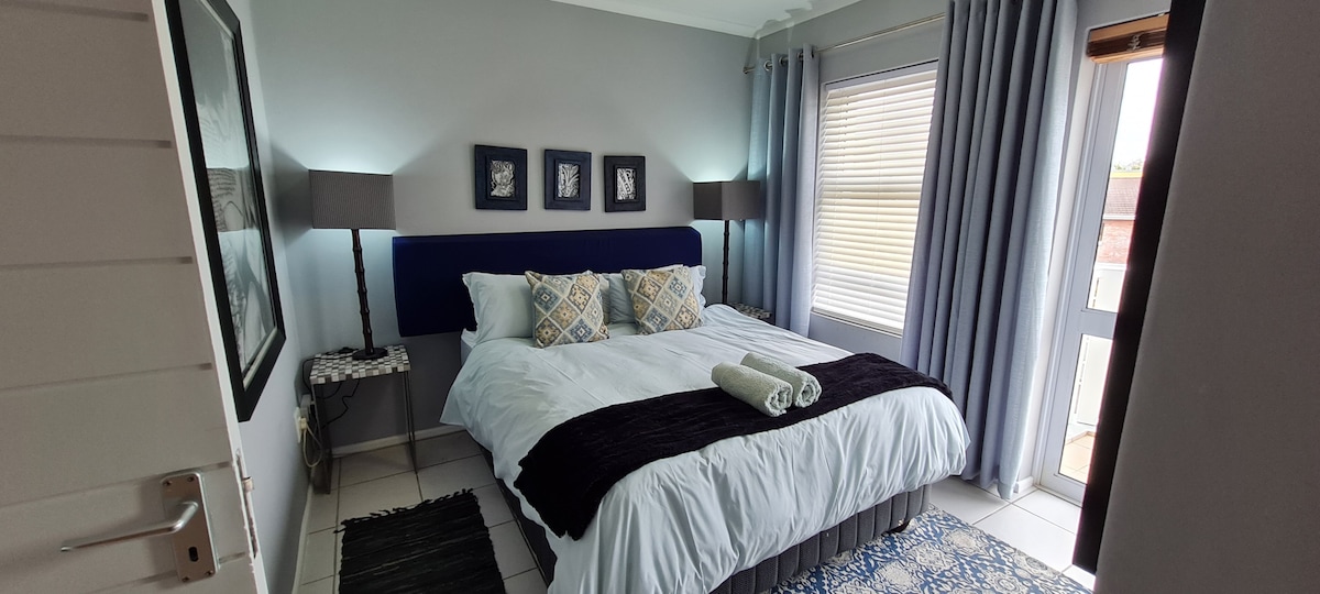 A serene bedroom is equipped with a spacious queen-size bed, adorned with decorative pillows and a soft throw. Natural light filters through sheer curtains, while two bedside lamps provide additional lighting. A door leads to an outdoor area, enhancing the room's bright atmosphere.