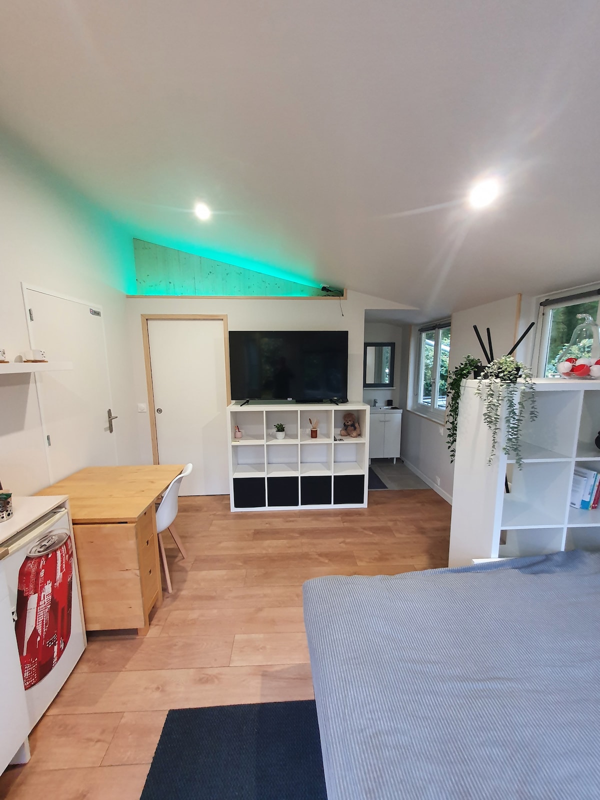A modern studio space is displayed, featuring a spacious bed with gray bedding. A wooden desk is positioned beside a white shelving unit, which holds decorative items. A large television is mounted on the wall, with natural light filtering through multiple windows, enhancing the overall ambiance.