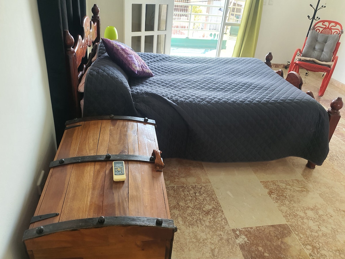 A spacious sleeping area features a dark quilted bedspread over a generous bed. A wooden chest with metal accents sits beside the bed, while a purple decorative pillow adds a pop of color. Natural light streams through a large window, partially covered by light curtains.