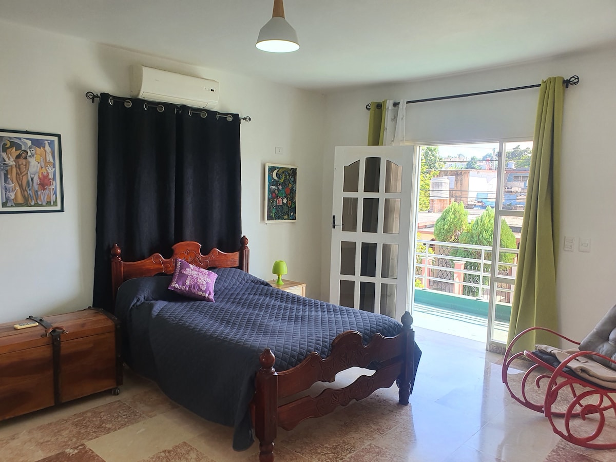 A spacious bedroom features a wooden bed with a dark blue quilt and a decorative pillow. Natural light enters through large glass doors leading to a balcony. A stylish red chair and a chest provide additional seating and storage.