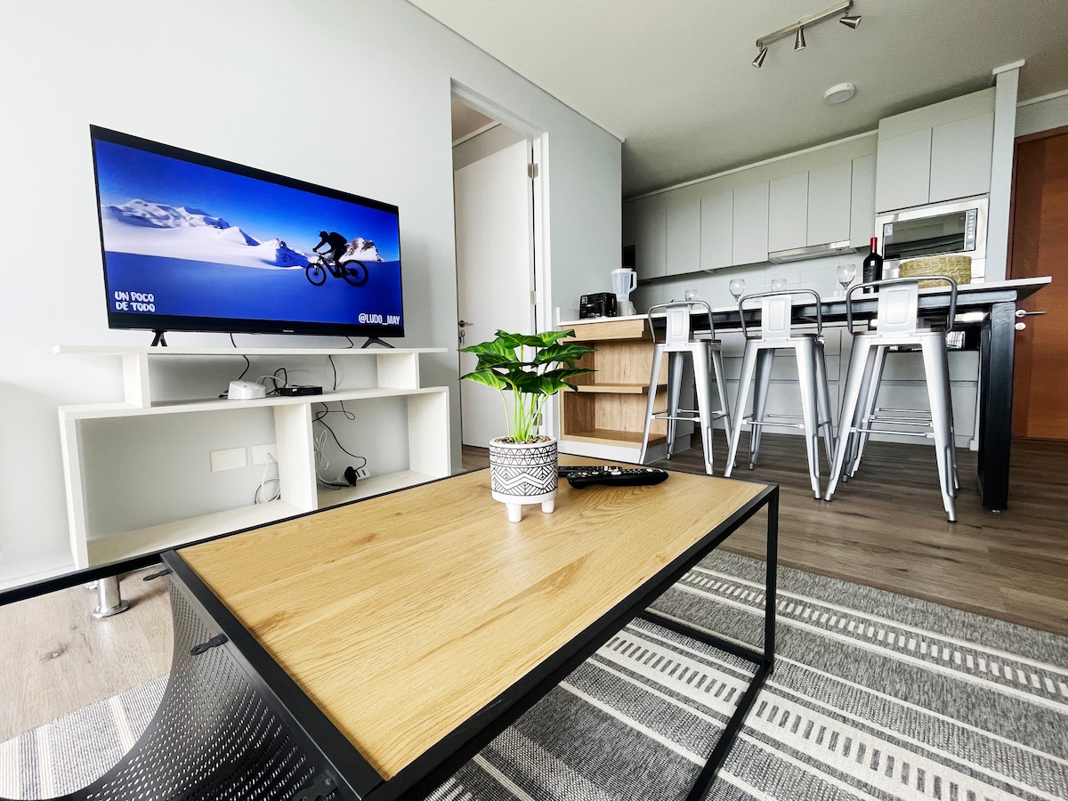 A modern living area features a flat-screen television atop a minimalist entertainment unit. A wooden coffee table sits in front, accompanied by a small potted plant. The open kitchen area is visible in the background, showcasing sleek cabinetry and high-backed bar stools.