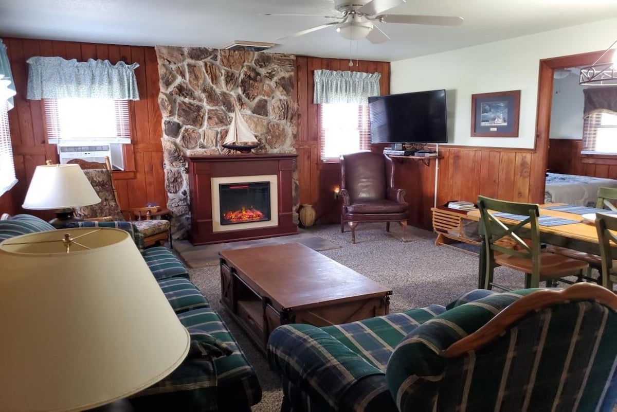 A cozy living room is equipped with a stone fireplace, complemented by a large flat-screen television. A plush couch and an armchair provide comfortable seating. Light filters in through window treatments, while a dining area with green chairs is visible in the background.