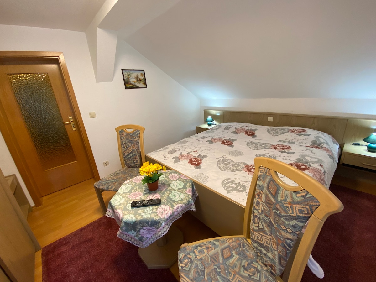 A spacious bedroom features a large bed with a floral-patterned coverlet. Two chairs with ornate backs flank a small round table adorned with a vase of flowers. Soft lighting from bedside lamps creates a warm and inviting ambiance.