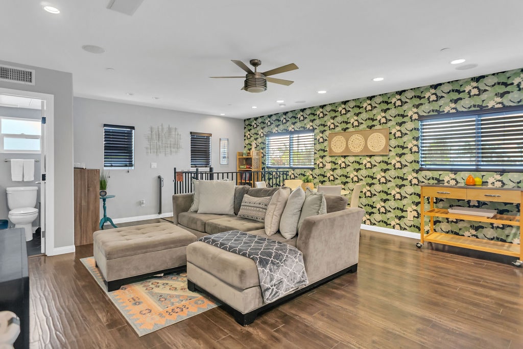 A spacious living area is illuminated by natural light through multiple windows. A large sectional sofa is positioned on a hardwood floor, complemented by decorative throw pillows. Unique wall art and a patterned accent wall add character to the room. A bar cart is visible in the corner.