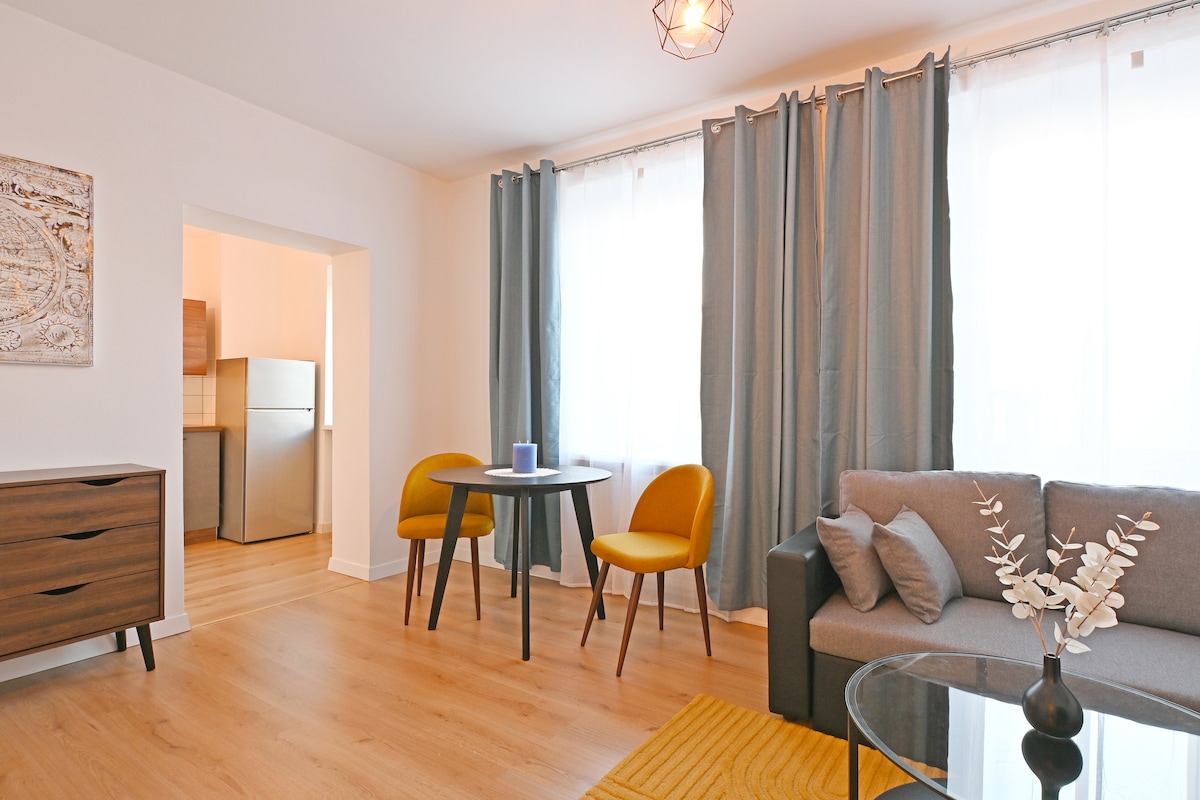 A light-filled living area features a grey sofa and a round glass coffee table. Two yellow chairs accompany a small dining table. A modern light fixture hangs from the ceiling, while sheer curtains frame the window, allowing soft natural light to enhance the space.