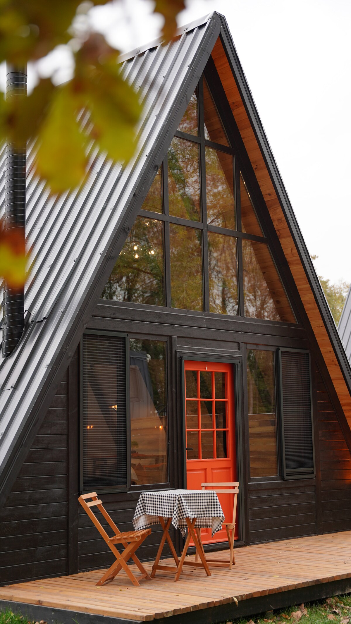 An A-frame cabin features a striking triangular design with large windows allowing for ample natural light. A small outdoor dining area with a checkered tablecloth is set with two wooden chairs, inviting moments of relaxation in the tranquil surroundings.