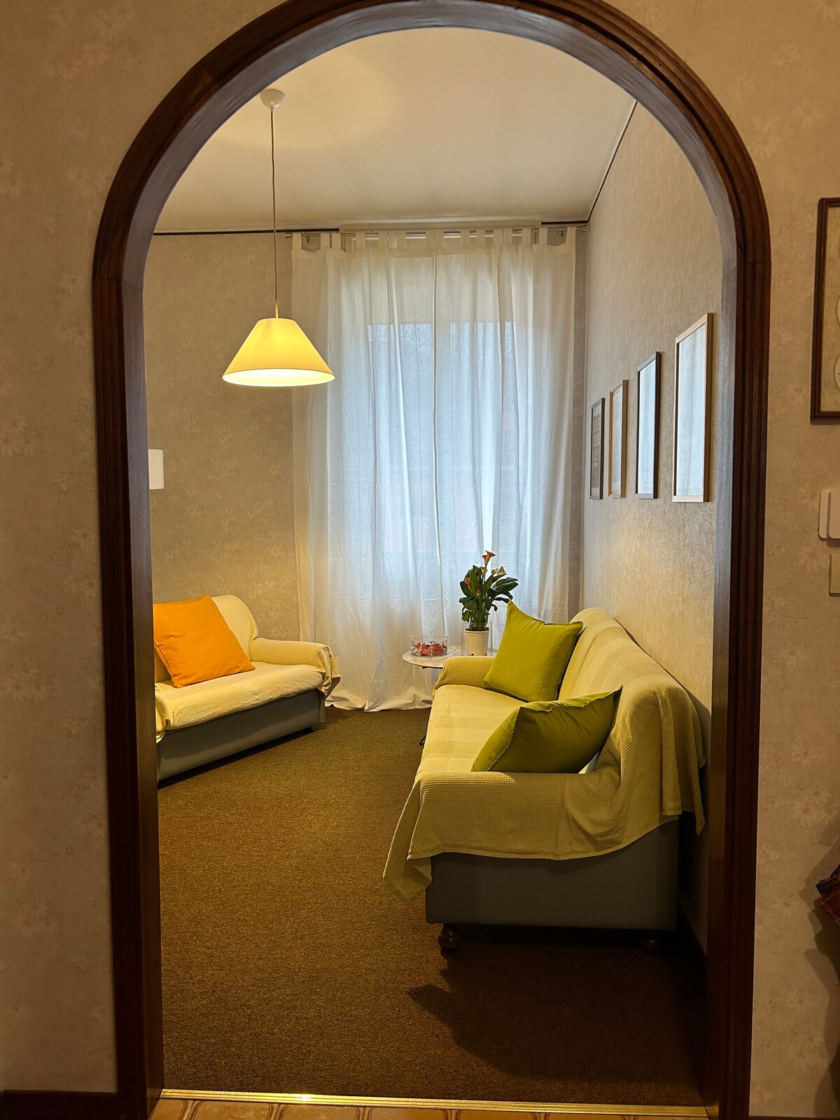 A welcoming living area is framed by an arched entrance. Soft natural light filters through sheer curtains, illuminating two sofas adorned with colorful cushions. A small table rests beside a potted plant, contributing to a relaxed ambiance.