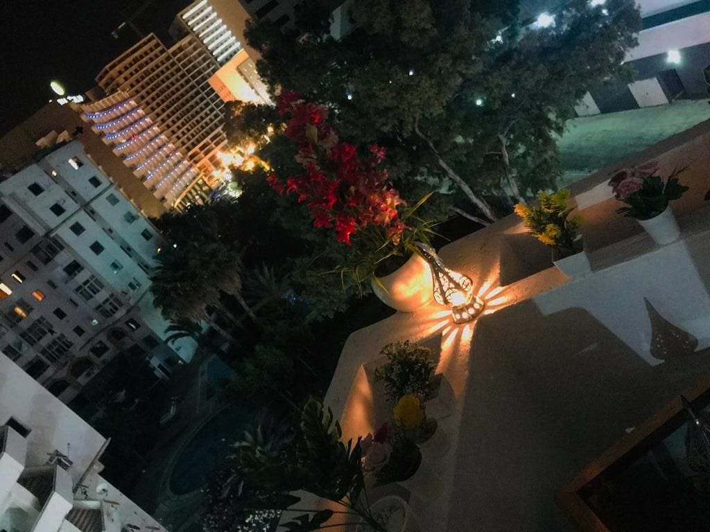 An illuminated balcony presents a view of the surrounding area at night. Floral arrangements are displayed on a white surface, casting shadows. In the background, city lights twinkle, including the outlines of buildings and palm trees, creating a serene atmosphere.