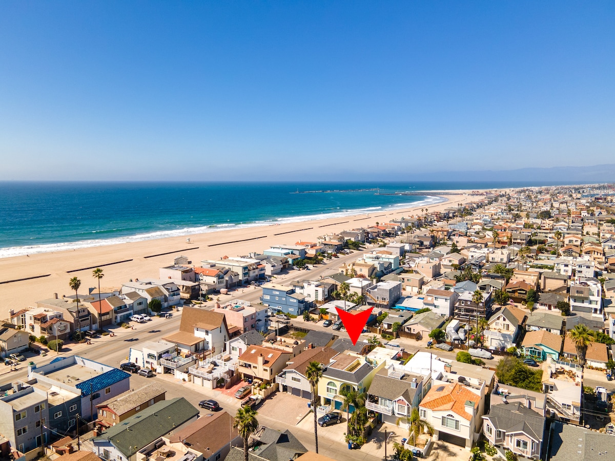 An aerial view captures the coastal area, with the beach stretching along the shoreline. The Seabreeze Cottage is highlighted, surrounded by other residential homes. The sparkling blue ocean reflects sunlight under a clear sky, enhancing the serene beach atmosphere.