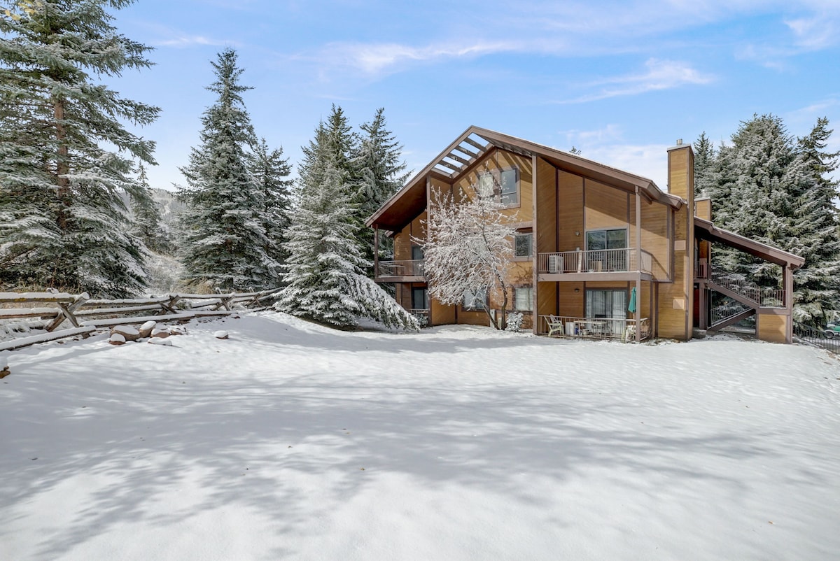 A cozy condominium is surrounded by snow-covered trees, with its three levels clearly visible. The rustic wooden exterior features large windows that allow natural light to fill the interior. An open area of snow is visible in the foreground, providing a tranquil winter setting.