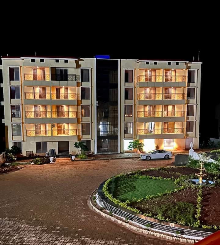 A modern building is illuminated at night, highlighting multiple balconies with extensive glass features. A landscaped garden surrounds the structure, featuring neatly arranged greenery and a decorative fountain in the foreground. A car is parked nearby, contributing to the welcoming atmosphere.