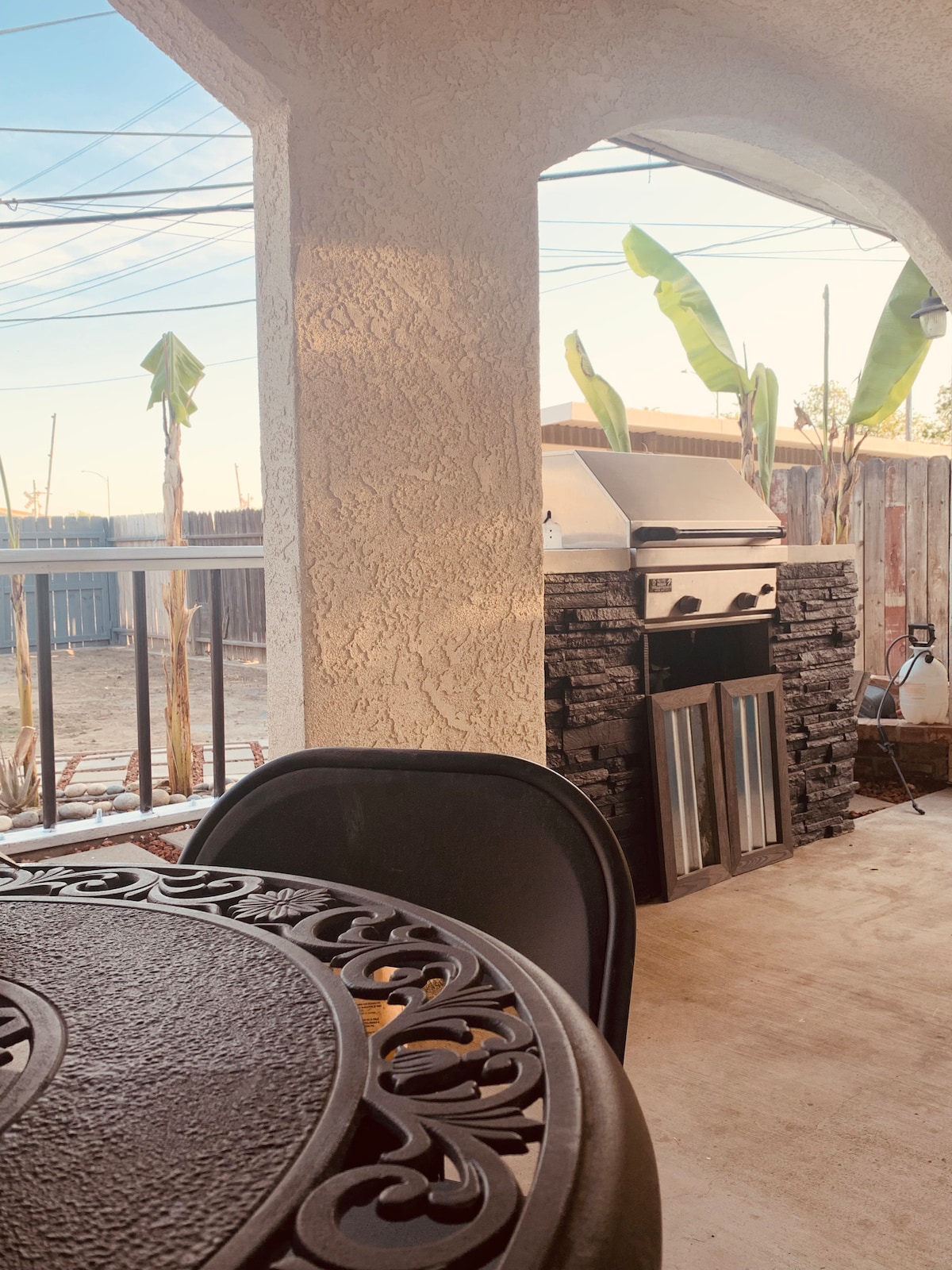 An outdoor patio area is depicted, featuring a sturdy table with intricate metalwork. A stainless steel grill is positioned nearby, situated against a stone backdrop. Sunlight casts gentle shadows while leafy plants stand tall in the background, creating a relaxed outdoor setting.