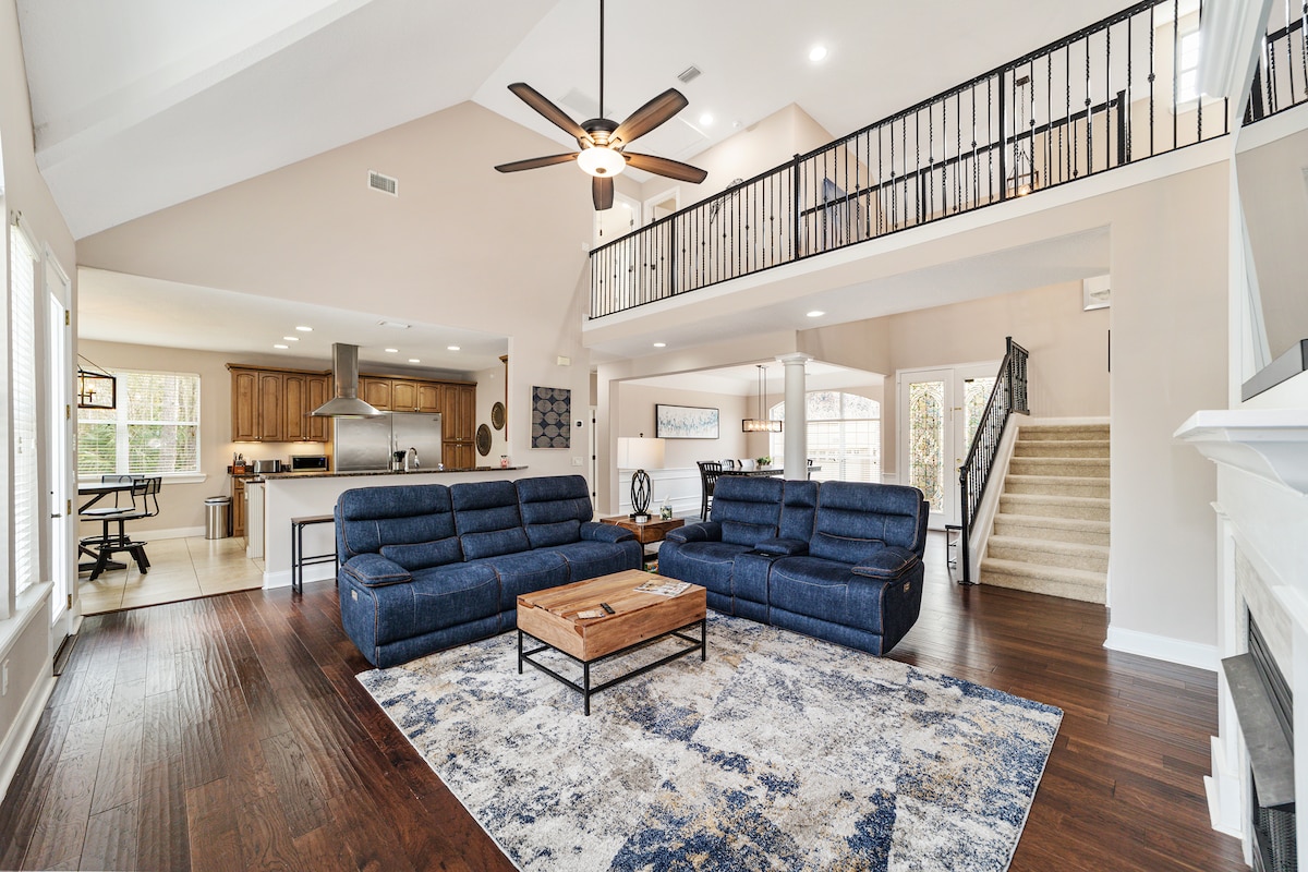 An open-concept living area is presented, featuring two spacious dark blue sofas facing a central coffee table. A large area rug with subtle patterns covers the floor. Natural light fills the room through multiple windows, with a staircase leading to an upper level and a modern kitchen visible in the background.