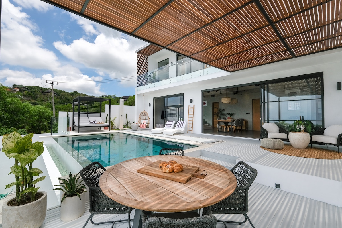An outdoor dining area features a round wooden table with a centerpiece of pastries. Surrounding the table are four textured chairs. The infinity pool is visible, along with comfortable sun loungers and a serene landscape in the background, complemented by a spacious outdoor patio.
