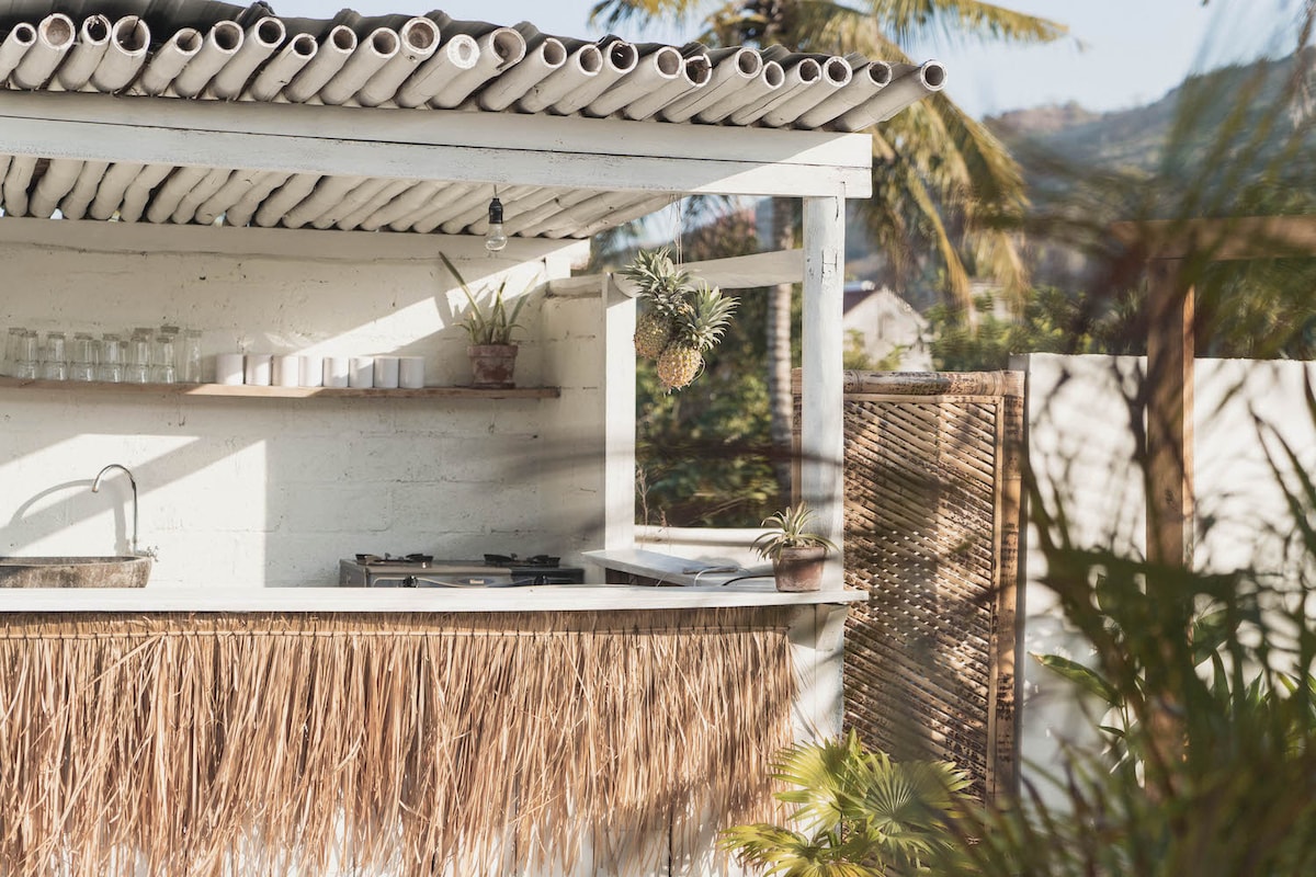 An outdoor kitchenette features a rustic wooden bar with a thatched design. Green plants are arranged on the countertop alongside glassware neatly placed on shelves. Natural sunlight enhances the inviting atmosphere, with palm trees partially visible in the background.