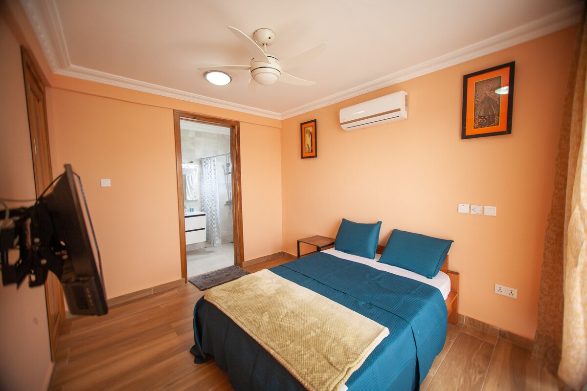 A spacious studio apartment features a bed with vibrant blue linens and a soft cream throw. Wall art is displayed on peach-colored walls, and a ceiling fan is installed above. A television is mounted at the foot of the bed, and natural light fills the room.