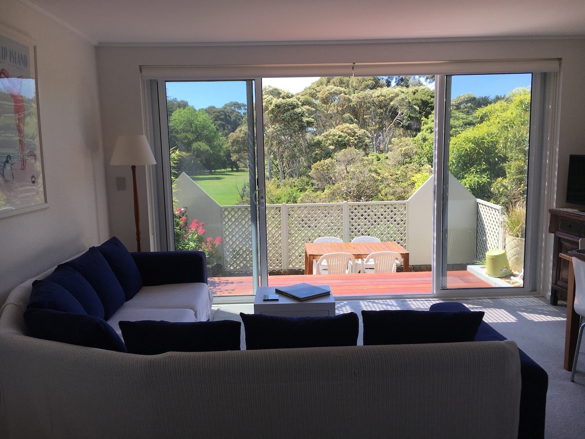 A spacious living area features large sliding glass doors that open to a deck surrounded by greenery. A comfortable sofa in blue and white provides seating, while a table with white chairs is positioned outside for dining. Natural light fills the room, enhancing the serene atmosphere.