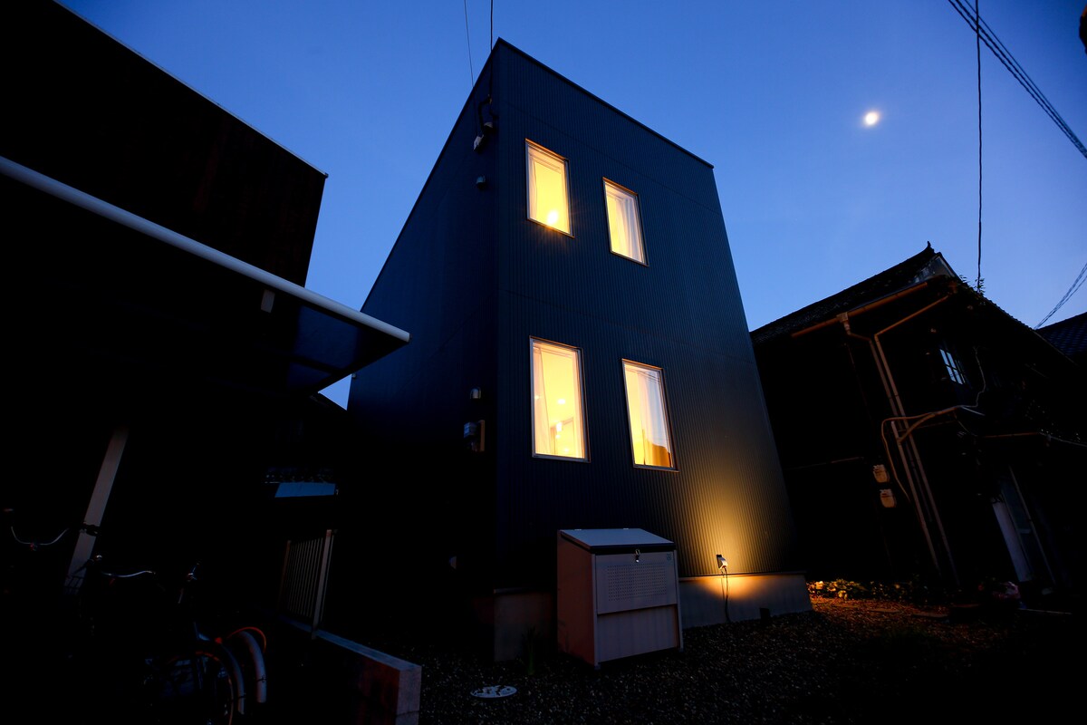 A modern, two-story building is shown at twilight, with illuminated windows reflecting a warm light. The exterior features a sleek dark facade, and a small storage shed is positioned in the foreground. The surroundings include gravel and nearby structures, creating a charming neighborhood setting.