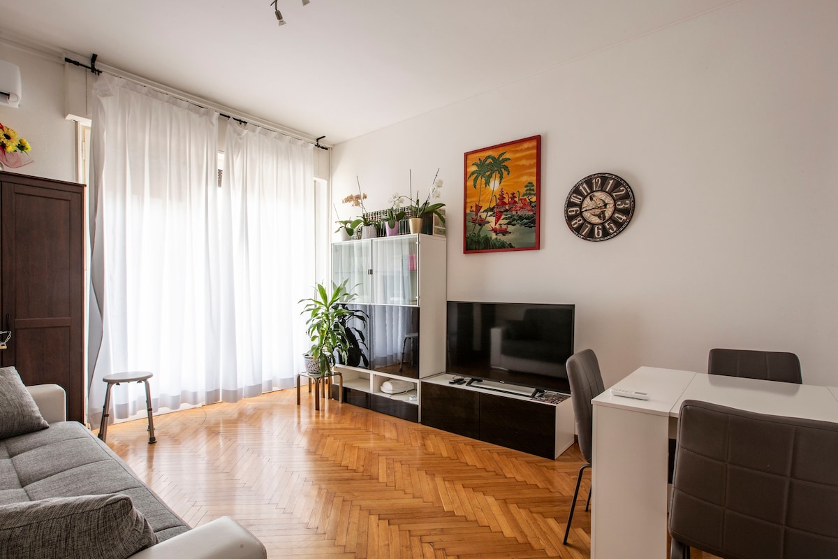 A well-lit living area features wooden flooring and large windows covered with sheer white curtains. A comfortable sofa sits adjacent to a wall-mounted smart TV, while a glass cabinet displays plants. A small dining table with chairs is placed to one side.